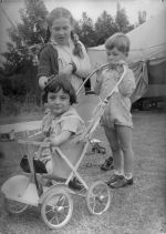 Long shot of three children. One child has an arm in a sling, one child is sitting in a pusher and the third child is holding the handle of the pusher. Playing Outside, Circa 1955