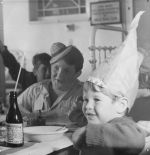 A medium shot of three children. Two are wearing party hats. There is a bed in the background on the right hand side and a bottle of lemonade with a straw in it and a bowl in the foreground on the left hand side. Celebrations at the Hospital, Circa 1955