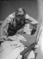 A medium shot of a female leaning over a young man, covered by a blanket, who is reclining in a reclining chair. He has his left hand covering her right hand. Enjoying the Sunshine, Circa 1955