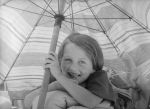 A close up shot of a young child lying on their stomach under a beach umbrella, smiling for the camera. Under the Sun Umbrella, Circa 1955
