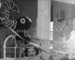 A medium shot of a young child smiling for the camera. The shot shows the child's head only. They are lying in a bed and the bed is decorated with streamers above it and there is a drawing of a fish on the wall behind the bed. Decorations at the Hospital, Circa 1955