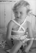 A medium shot of a young child sitting in a cot, wearing a harness. The child is holding a pencil in their left hand and appear to be holding a writing pad in their right hand. Portrait of a Child, Circa 1955