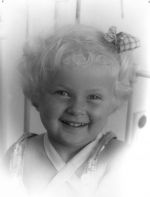 A close up shot of a young child smiling for the camera. There are the bars of bed behind them. Portrait of a Child, Circa 1955