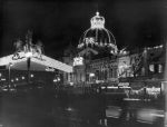 A medium shot taken at night of a street, with buildings in the background and two people in the foreground looing toward the buildings. Above the buildings is a crown made of lights, with ER lit up in the middle. In the foreground, on the left hand side, is a structure whose roof is made of lights, with a crest on the corner facing the camera. Welcoming Queen Elizabeth II, Circa 1955