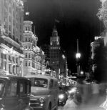 A medium shot of a street, showing buildings and cars. It is taken at night, some of the cars and buildings have lights on and the street lights are on. Welcoming Queen Elizabeth II, Circa 1955