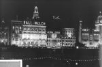 A long shot showing a number of buildings along Flinders Street, Melbourne, Victoria. The photograph is taken at night and the buildings have lights on their facades and some have their names in lights - Ball & Welch, Majestic, and State Theatre. Welcoming Queen Elizabeth II, Circa 1955
