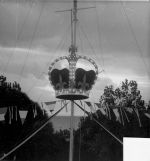 A medium shot, taken outside, of a model of a crown, suspended on a pole, with streamers radiating from below it. The crown is lit with strings of lights. Welcoming Queen Elizabeth II, Circa 1955