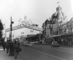 A long shot showing a street and buildings decorated for a visit by royalty. One of the buildings is topped with a crown and there is a structure spanning the street, decorated with the royal crest. There are cars on the road and people walking on the footpaths. Welcoming Queen Elizabeth II, Circa 1955