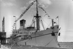 A long shot of a ship alongside a wharf. The ship has bunting extending up across the masts from the bow to the stern. The word 'GOTHIC' is visible on the right side of the ship near the bow. Ship in Dock, Circa 1955