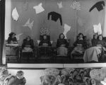 A long shot of seven people on a stage. The person on the far left is lying on a bed facing the camera; the next four people are sitting in wheelchairs facing the camera with blankets on their knees; the fifth person is sitting facing the camera although partially obscured by the sixth person in a wheelchair with a blanket on her knees. In front of the stage is a small chair and a couch. A young child is sitting on the chair looking back at the stage. Behind the stage is a curtain with items of clothing attached to it. Patients on Stage, Circa 1955