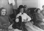 A medium shot of five people seated looking at the camera. Three in front are seated in wheelchairs; two of whom are wearing hats. A woman is seated slightly behind the second and third person and another person is just visible on the right hand side of the photograph. Patients on Stage, Circa 1955