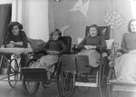 A medium shot of four people looking at the camera; first person is lying on a bed and next three are sitting in wheelchairs. The fourth person is only half visible in the photograph. Patients on Stage, Circa 1955