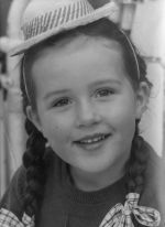 A close up shot of a young child looking at the camera. Child has pigtails with ribbons at the ends and is wearing a small hat held in place by elastic. Portrait of a Child, Circa 1955
