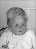A close up shot of a young child wearing a harness, looking slightly off camera. The bars of a bed are visible behind the child. Portrait of a Child, Circa 1955