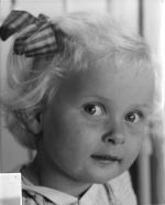 A close up shot of a young child looking at the camera. Child has short fair hair tied at the side with a bow. Portrait of a Child, Circa 1955