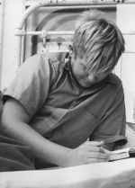 A close up shot of a young man leaning on his left elbow on a bed looking down at an open book. The bars of the bed are visible behind him. Patient in Bed, Circa 1955