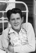 A close up shot of a young man looking at the camera. He is holding his left arm across his body. The bars of the bed are visible behind him. Patient in Bed, Circa 1955