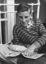 A close up shot of a young man looking at the camera. He is leaning on his right elbow with his left hand on an open comic book. The bars of the bed are visible behind him. Patient in Bed, Circa 1955