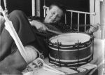 A close up shot of a young man lying back on a bed. He has a drum on the bed beside him and is holding a drumstick in each hand. A newspaper is visible under the drum. Left leg appears to be in traction. The bars of the bed are visible behind him. Playing Musical Instruments, Circa 1955