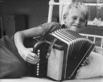 A close up shot of a young child looking at the camera. Child is leaning on left elbow on a bed holding a piano accordion. The bars of the bed are visible behind him. Playing Musical Instruments, Circa 1955