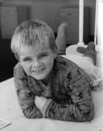 A close up shot of a young child lying on a bed, smiling at the camera. The bars of the bed and two small cabinets are visible behind the child. Portrait of a Child, Circa 1955