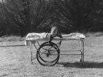 A medium shot of a young child lying outside on a bed. Bed is a trolley-style with two large wheels. Child is looking at the camera with one hand on one of the wheels. Trees are visible in the background. Young Child on Trolley, Circa 1955