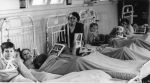 A medium shot of a woman sitting in between hospital beds. Six children are visible: first is sitting up in bed wearing a harness, second is lying in bed, third is slightly obscured but is holding a picture, fourth is leaning back in bed, fifth is sitting between two beds and sixth is lying in bed. Reading in Bed, Circa 1955