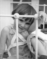 Medium shot of a child seated on a bed one arm holding the bed rail with braces fitted over shoulder and chest posing for the camera. Portrait of a Child, Circa 1955