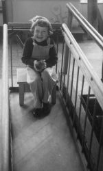 A medium shot of a young child sitting on a stool holding something in both hands. Stool is positioned in between two railings. Portrait of a Child, Circa 1955