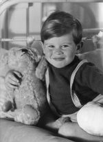 A close up shot of a young child sitting on a bed with his right arm around a teddy bear. Right leg appears to be in plaster. Portrait of a Child, Circa 1955