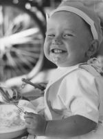 A close up shot of a young child wearing a hat smiling at the camera. Child is holding a spoon in their left hand and something in their right hand and has food on their face. A bowl is visible in front of the child. Portrait of a Child, Circa 1955
