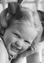A close up shot of a young child looking at the camera. Child has short fair hair tied at both sides with ribbons and is leaning on left hand. Portrait of a Child, Circa 1955
