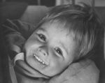 A close up shot of a young child smiling at the camera. Child is lying back with arms behind their head. A harness is partially visible. Portrait of a Child, Circa 1955