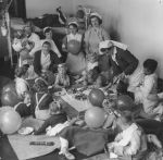 A long shot of a party with multiple nurses and children, party food, balloons visible. A hospital bed is visible in the background. Birthday Celebrations at the Hospital, Circa 1955