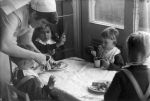 A medium shot of a nurse leaning over a table feeding a young child in front of her from a cup. Three other young children are seated at the table eating. Dinner Time, Circa 1955