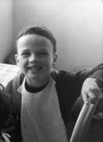 A close up shot of a young child seated at a table smiling at the camera. Child is wearing a bib and is leaning with left arm on the back of a chair. Portrait of a Child, Circa 1955