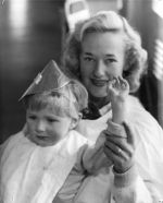 A close up shot of a woman sitting with a young child on her lap. She is holding the child's left arm up to wave to the camera. Child is wearing a bib and a party hat. Jenny Howard and Child, Circa 1955