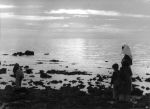 A long shot of a nurse and several young children standing on the beach near the shallows. Child on left of photograph is leaning over looking into the water. At the Beach, Circa 1955