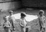 Medium shot of four children playing outside with a ball and sticks, a blanket placed on the grass behind them, a small wall in the background. Children Playing, Circa 1955