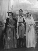 Long shot of two women and one man dressed in costumes. The woman on the left is carrying a wand, the woman on the right is wearing a white dress with veil, and carrying flowers. The man, standing between the women, is wearing a cape with fur trim. Pantomime at the Hospital, Circa 1955