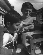 Medium shot of two children standing looking at a small wooden loom, one child has a brace fitted over shoulder and chest posing for the camera. Weaving on the Loom, Circa 1955