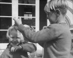 Medium shot of two children outside one child is standing placing ribbons on the seated child's hair, posing for the camera, a window in the background. Playing, Circa 1955