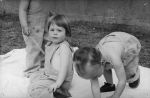Medium shot of three children on a blanket one child kneeling, the other is bending over from the waist with arms placed on the blanket, one child standing upper body is obscured due to the angle of the photograph holding a stick posing for the camera. Playing Outside, Circa 1955