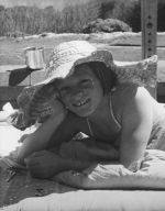 Medium shot of a child outside laying prone on a mattress wearing a broad brimmed hat both arms bent at the elbow one holding the head, metal cup behind child, with trees in the background. Portrait of a Child, Circa 1955