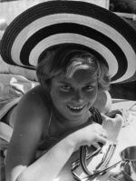 Medium shot of a young person outside laying prone on a mattress wearing a broad brimmed hat, metal cup in the foreground. Portrait, Circa 1955
