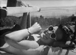 Medium shot of a young person outside laying supine on a mattress underneath a wooden shelf wearing sunglasses watch posing for the camera. Weaving on the Loom, Circa 1955