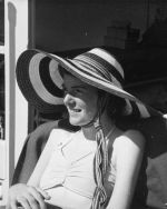 Medium shot of a young person seated outside wearing a broad brimmed hat posing for the camera. Sunhats on the Balcony, Circa 1955