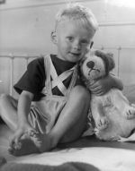 Medium shot of a child seated on a bed legs bent at the knee, one hand holding his feet the other hand holding a stuffed toy posing for the camera. Child and Teddy Bear, Circa 1955