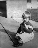 Medium shot of a child seated outside on a paved area with a toy in one hand. Brick building in the background. Sitting Outside, Circa 1955