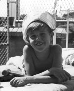 Medium shot of a child outside laying prone on a bed arms crossed at elbow wearing a hat a wooden mallet on the bed, posing for the camera, wire fence and a brick wall in the background. In the Sunshine, Circa 1955